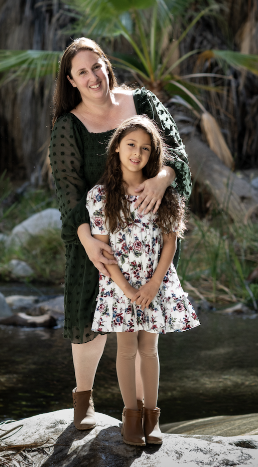 An outdoor portrait of a person in a green dotted dress standing with a child wearing a white floral dress, positioned on rocks with palm plants in the background.