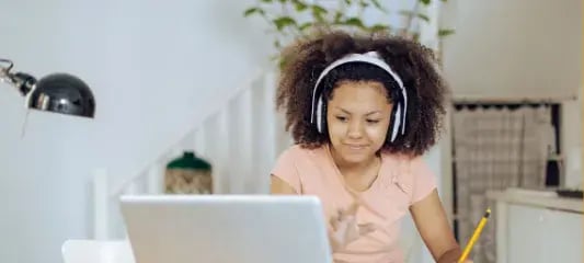 A young girl with curly hair wearing white headphones and a light pink top sitting at a laptop. She appears to be focused on her screen, possibly participating in online learning. There's a pencil visible and the setting looks like a home study space with plants and a lamp in the background