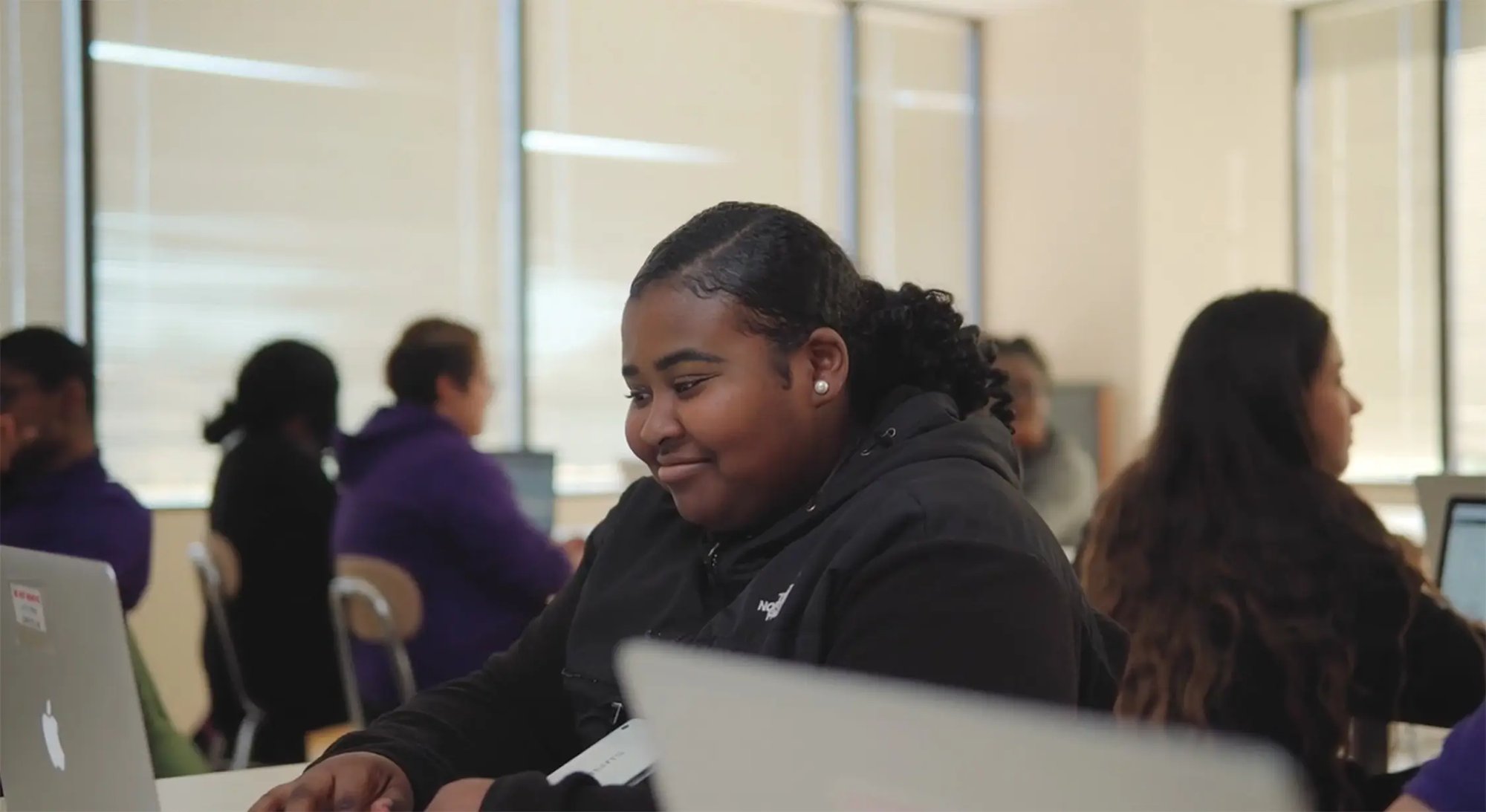 A young student with dark hair styled in a ponytail sits at a computer in a classroom setting. They are wearing a black hoodie and appear focused on their work. Other students can be seen in the background at computer workstations.