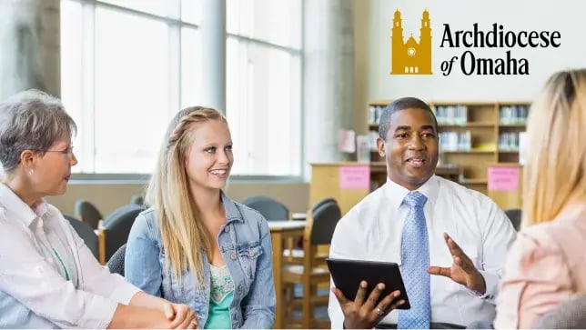 A diverse group of four educators in a meeting with the Archdiocese of Omaha logo visible in the top right corner. A man in a white shirt and blue tie is holding a tablet while gesturing as he speaks to three colleagues in a bright library or resource room. A diverse group of four educators in a meeting with the Archdiocese of Omaha logo visible in the top right corner. A man in a white shirt and blue tie is holding a tablet while gesturing as he speaks to three colleagues in a bright library or resource room.