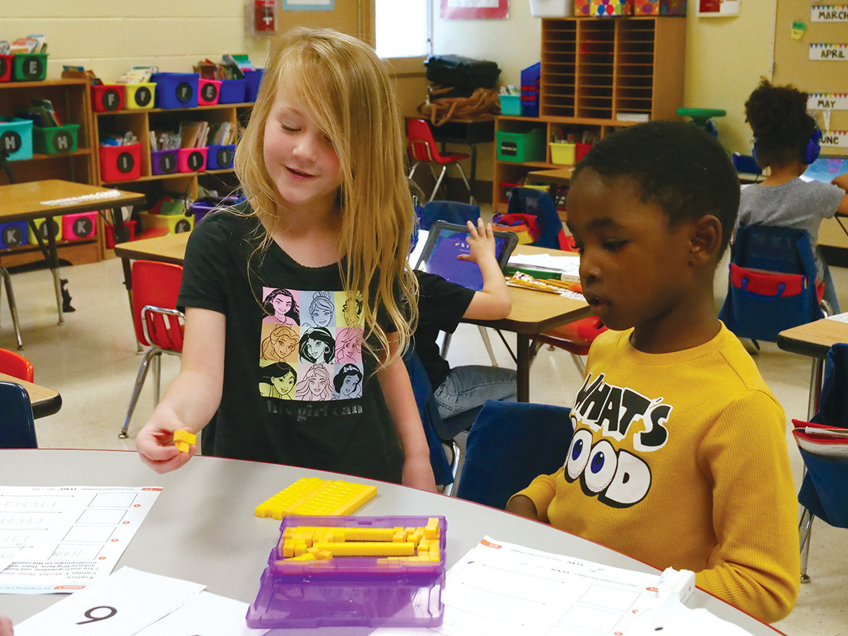 Two elementary students work together at a desk using yellow math manipulatives from a purple container. 