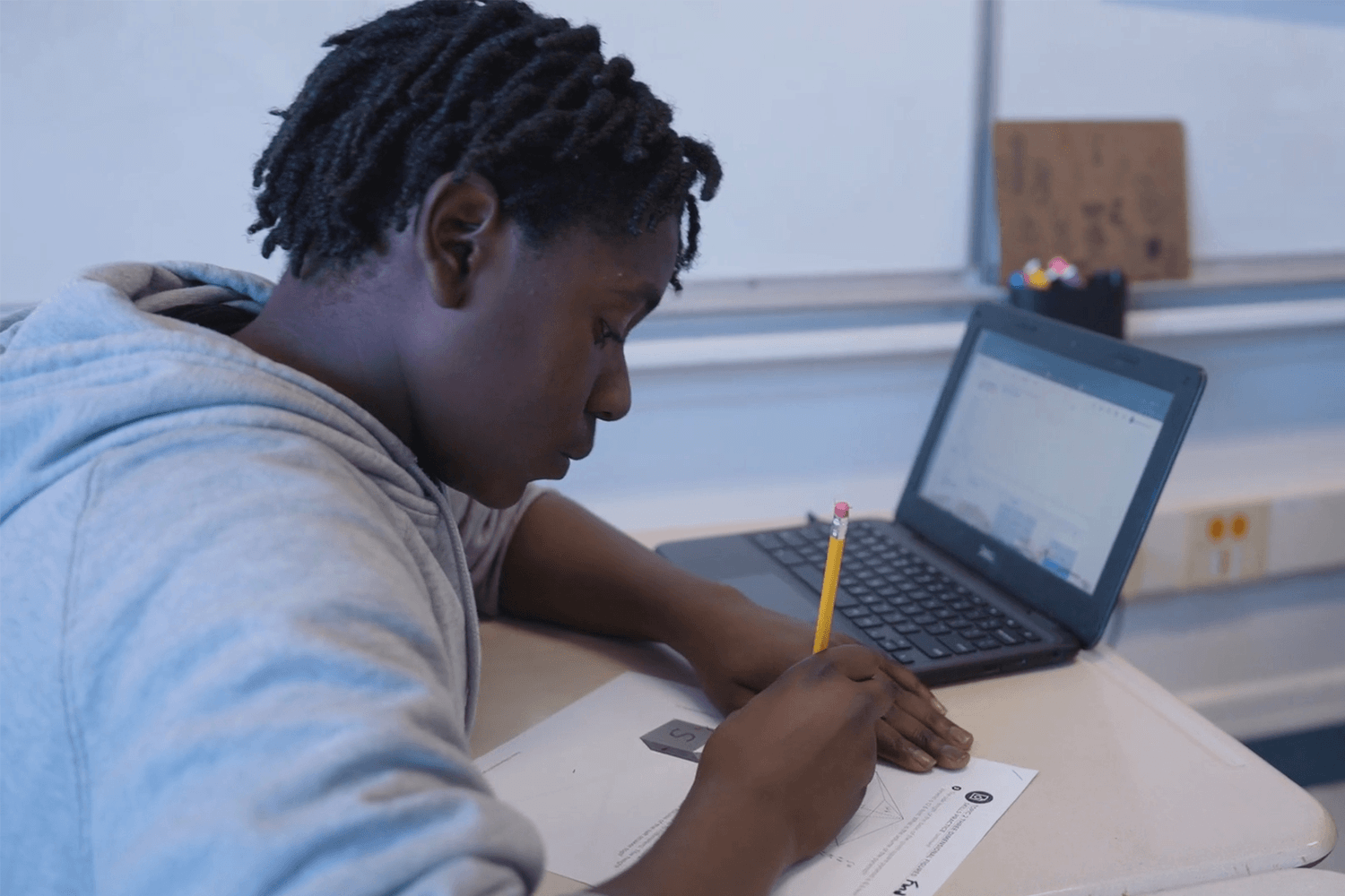 A student with short dark curly hair works at a classroom desk, writing on a math worksheet with a pencil and a laptop