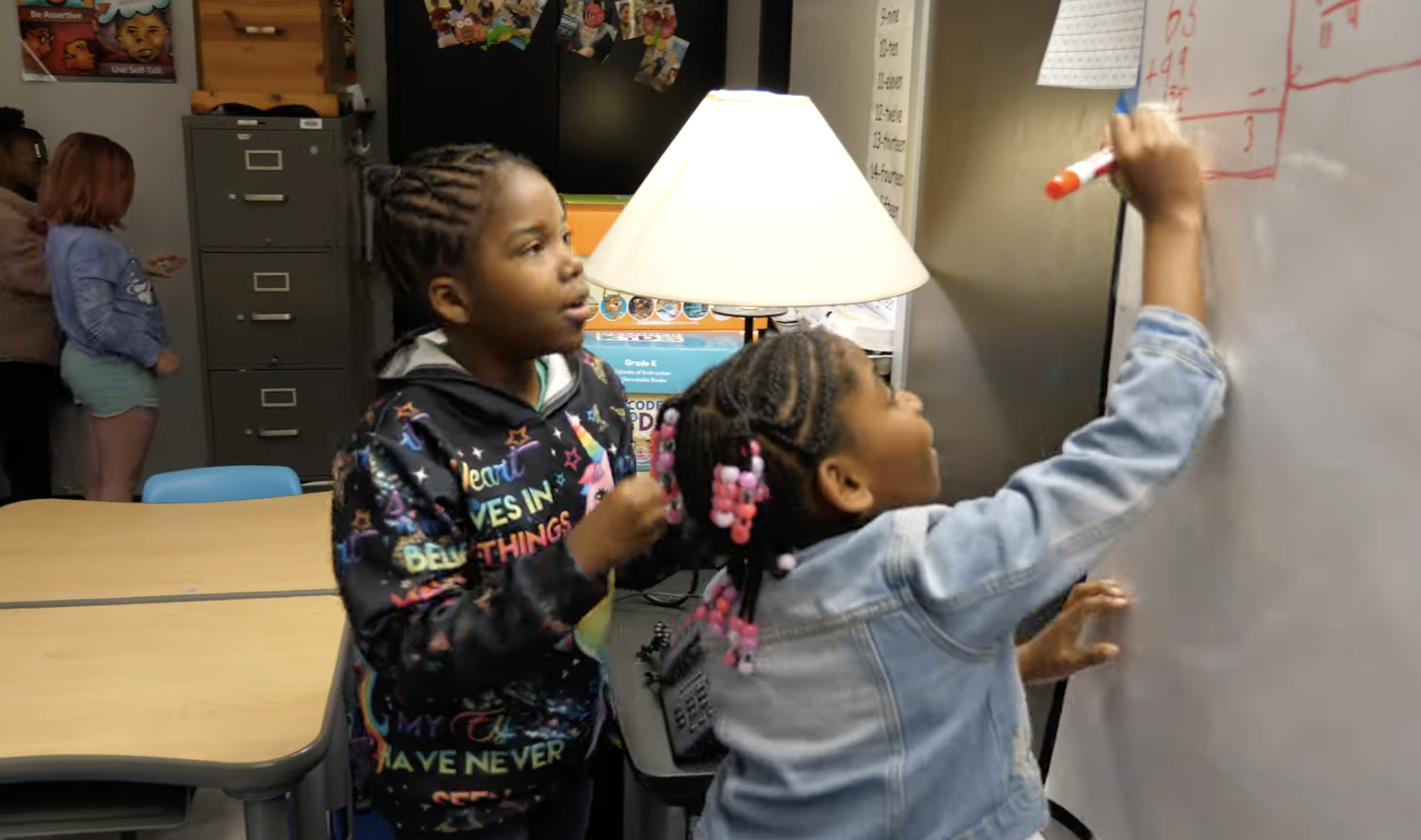 Two elementary students solve an addition problem together at a classroom whiteboard.