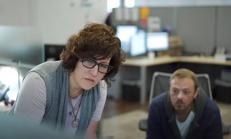 A woman with curly hair and glasses leans forward, focused on something offscreen. Beside her, a bearded man mirrors her intensity. Theyre in an office, possibly diving into Carnegie Learning Research, surrounded by desks and monitors that reflect their dedication.
