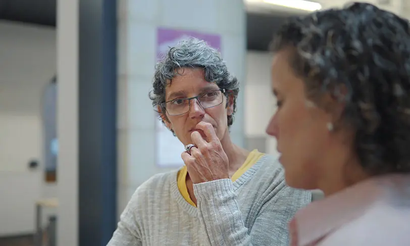 Two people are in conversation indoors. One, wearing glasses and a light-colored sweater with a yellow collar, looks intently at their companion, perhaps discussing Carnegie Learning Research findings. The other person is turned slightly away, deep in thought.
