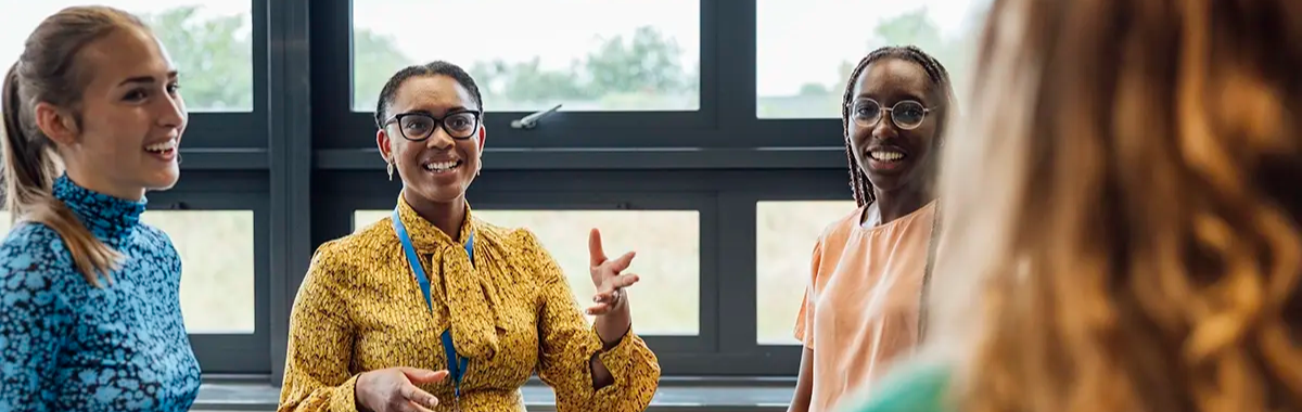 Three smiling educators engaged in conversation in a bright classroom setting.