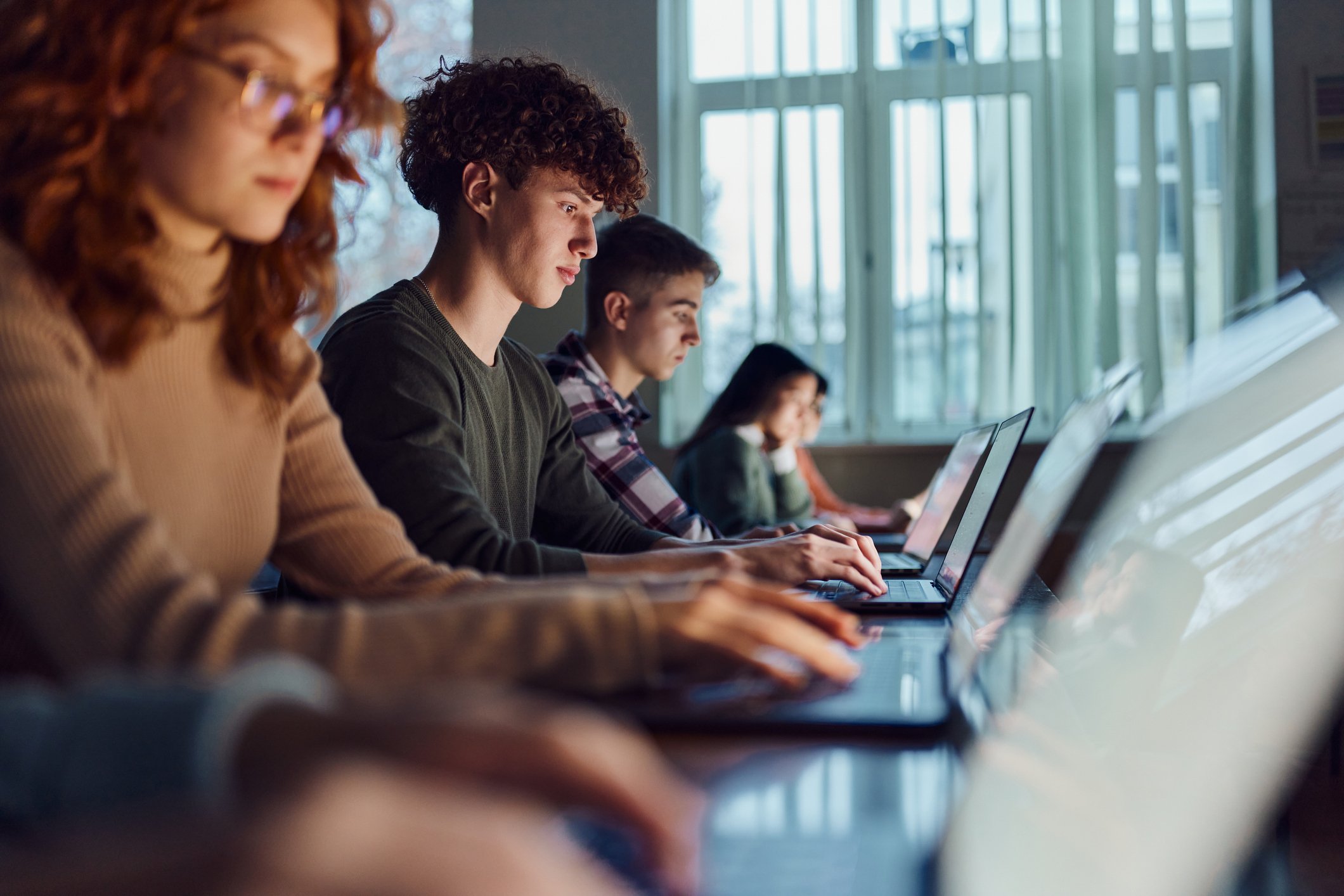A group of students sit in a row at desks, focused on typing on their laptops in a well-lit classroom with large windows.
