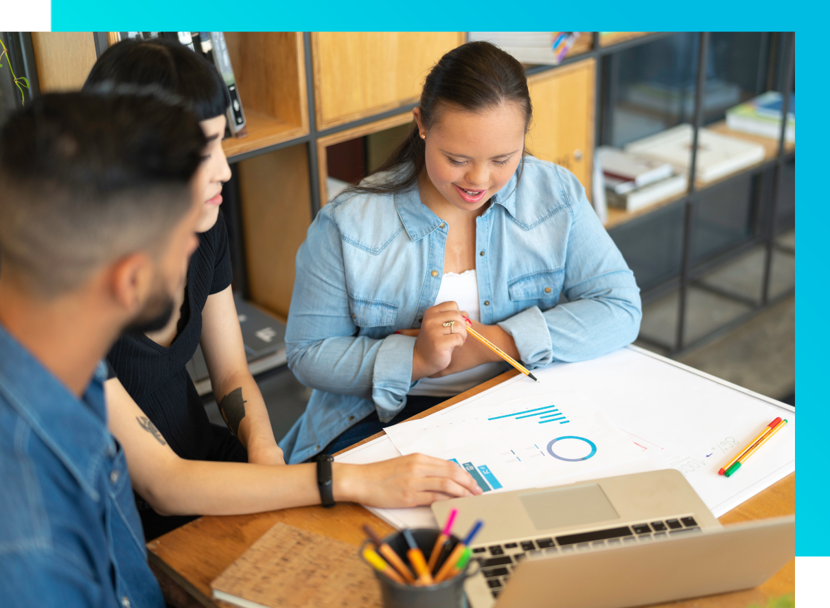 Three people seated around a table in a modern workspace, collaborating on a project. One person in a light blue shirt is looking down at documents with charts or graphs. Another person is pointing at the paperwork, which appears to contain colorful data visualizations or infographics. 