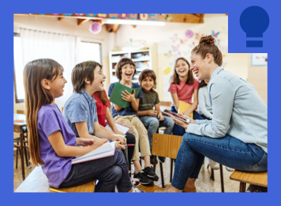 An adult with light-colored clothing sits in a chair reading a book to a small group of children who are seated in a semi-circle. The children appear to be elementary school age and are listening attentively in what looks like a classroom or library setting. 