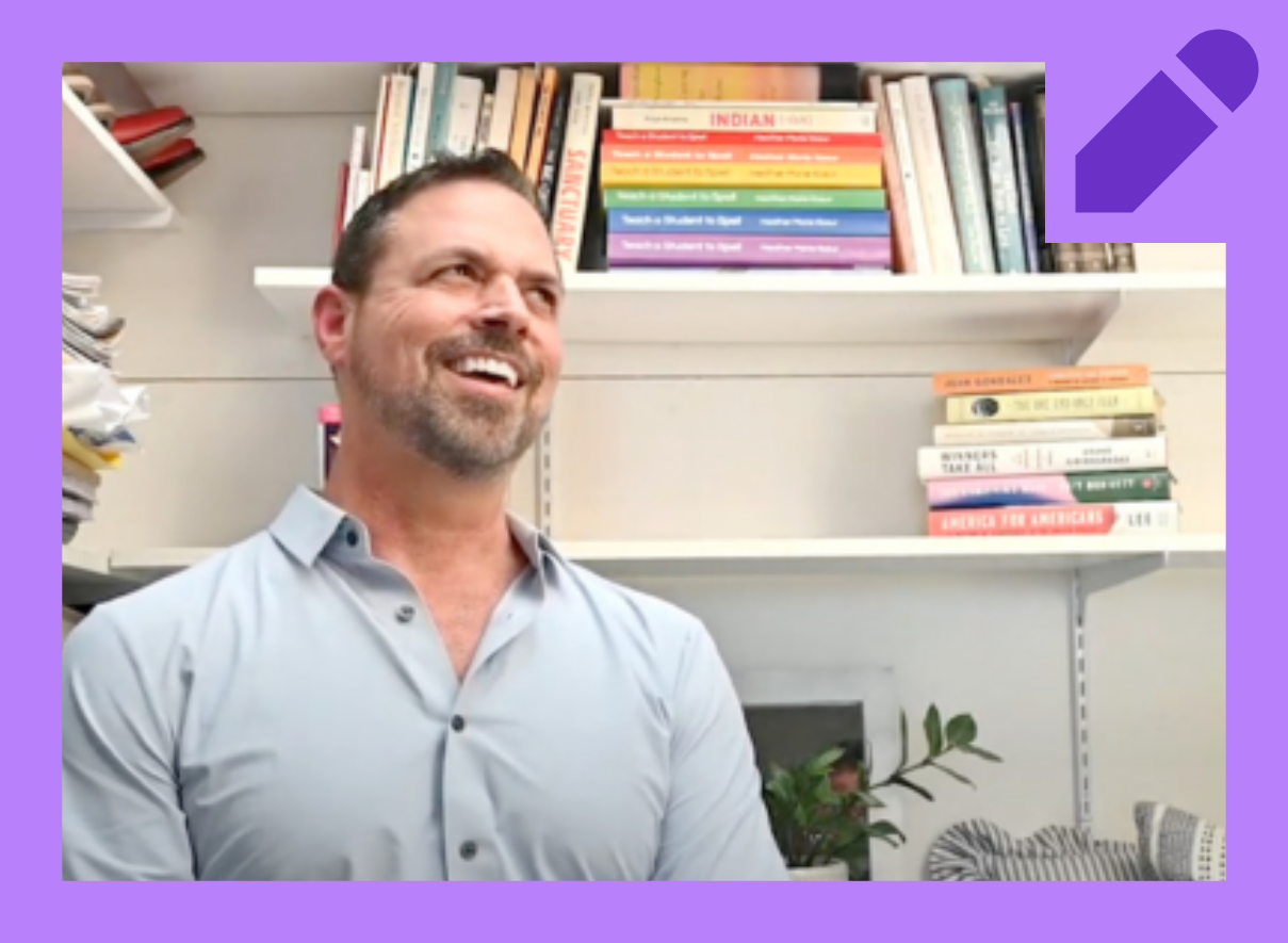 A smiling man with facial stubble wearing a light blue button-up shirt. He appears to be in an interior space with white shelving or cabinets visible in the background.