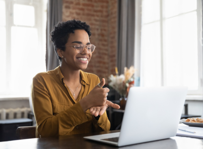 Smiling Black woman with natural hair working on a laptop in a casual setting with brick walls visible in the background. She wears a brown top and appears engaged with something on her screen.