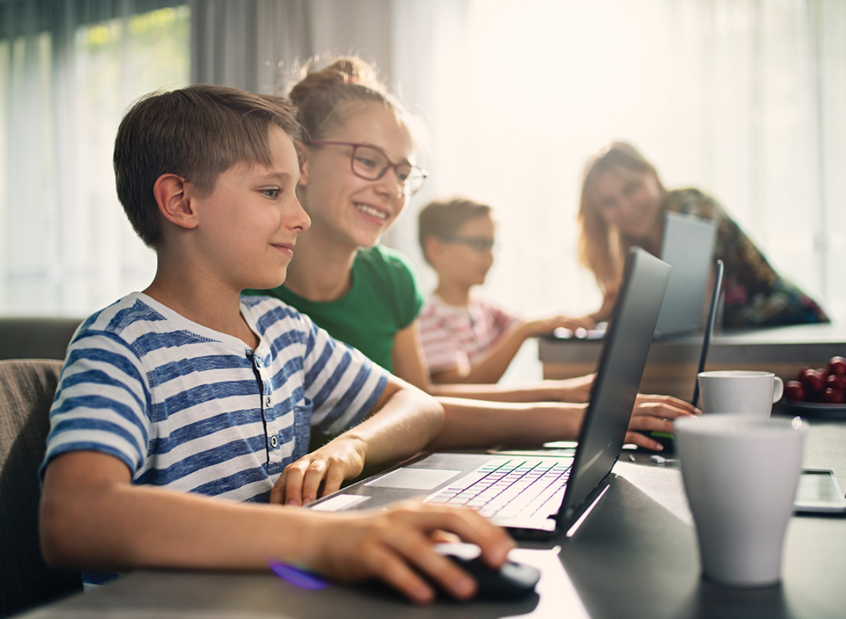 Students working at computers in a classroom. In the foreground, two students sit side by side at desktop computers, with one wearing a striped shirt. In the background, additional students can be seen working at their own computers.