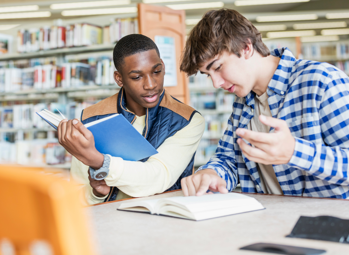 Two young men studying together in a library. One black student is holding a tablet or digital device, while a white student with light brown hair is taking notes in a notebook. 
