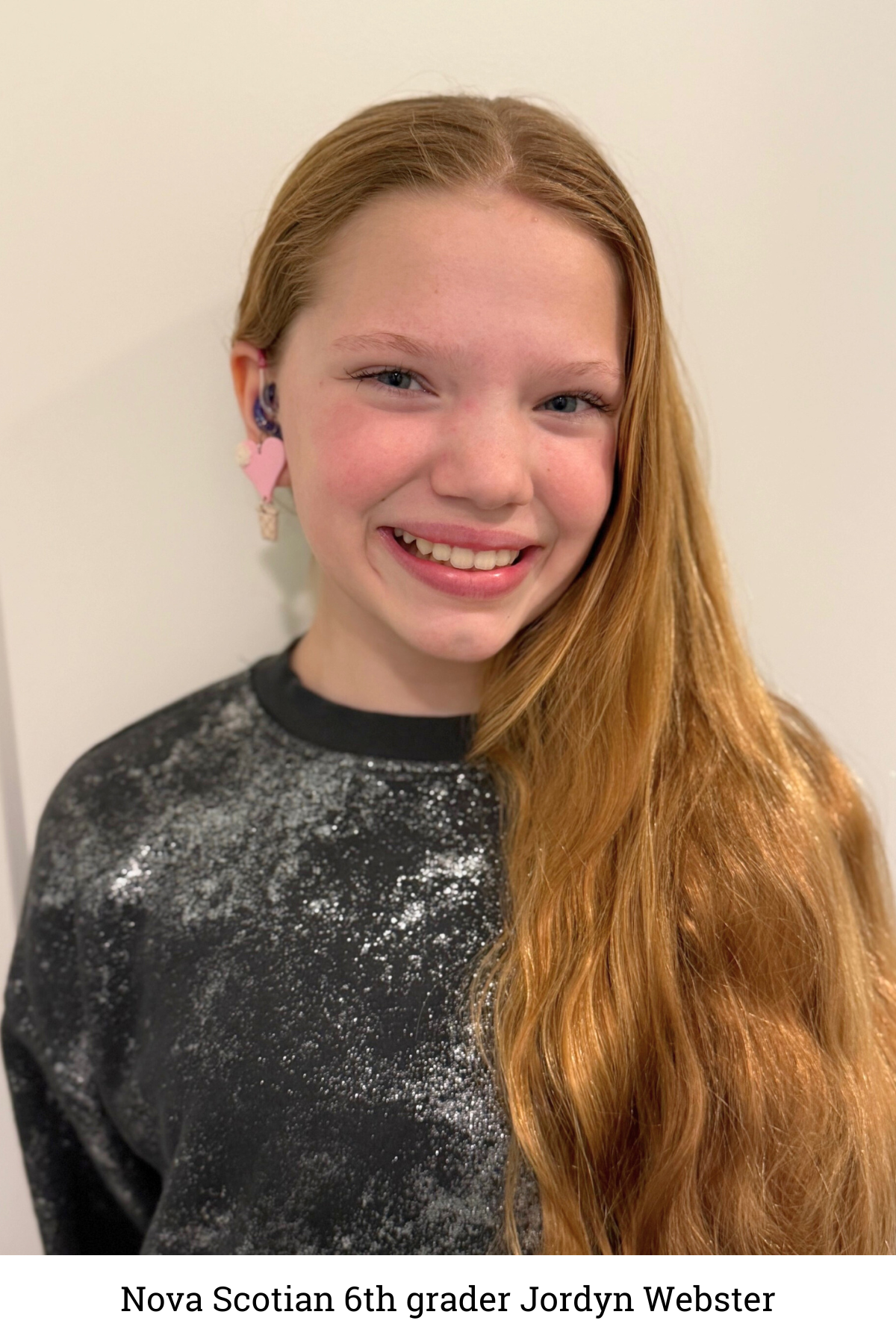 Sixth-grade student Jordyn Webster pictured with long, light brown hair, a black speckled shirt and pink heart-shaped earrings. She stands against a plain, light-colored background and smiles at the camera.