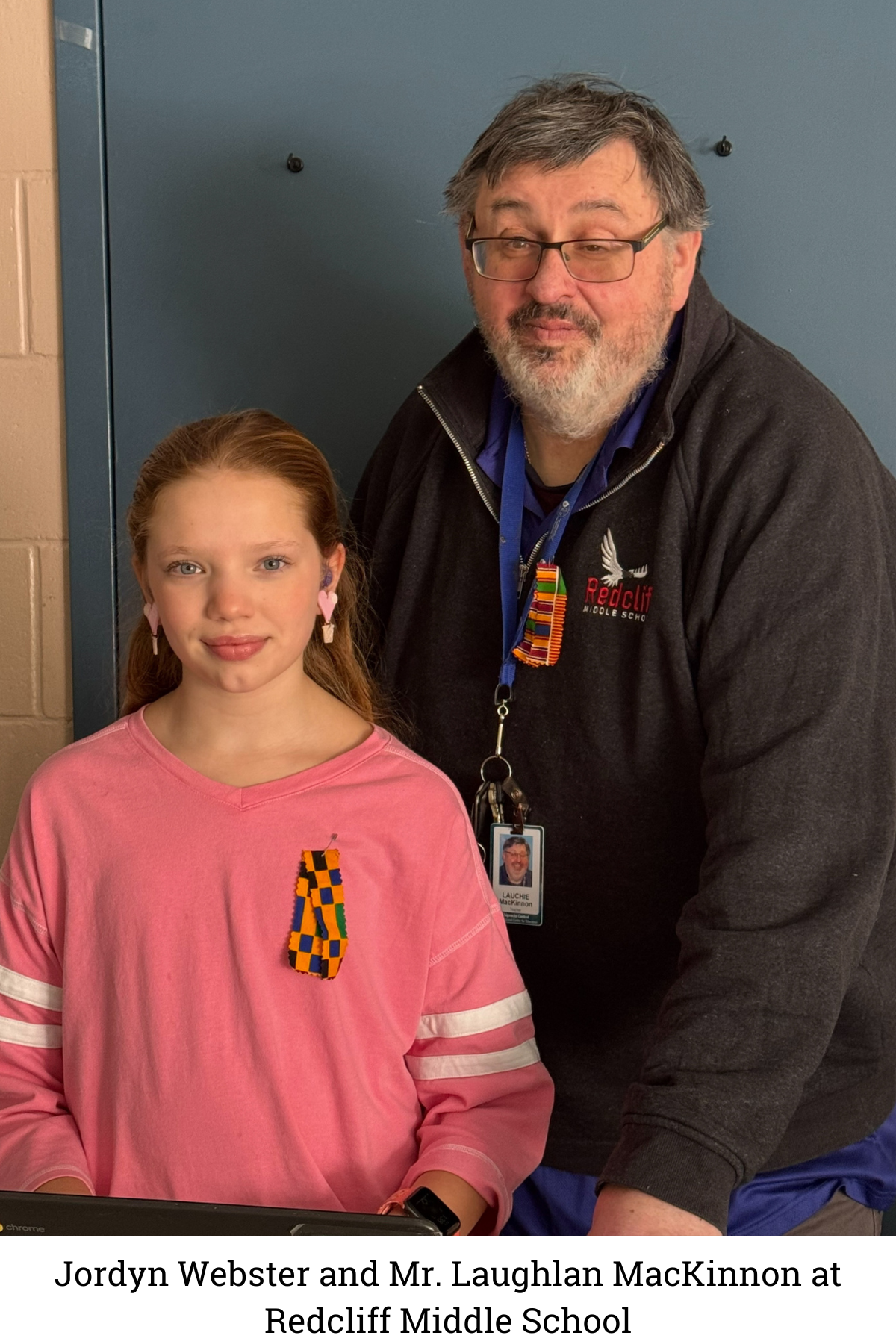 Jordyn Webster (pictured in a pink shirt) stands next to her reading intervention teacher, Mr. Laughlan MacKinnon, who is pictured wearing glasses with a beard wearing a black jacket. Both are smiling in a classroom at Redcliff Middle School in Nova Scotia, Canada. Jordyn gained 3 years of reading progress in 8 months working with Mr. MacKinnon and Carnegie Learning's Fast ForWord reading program.