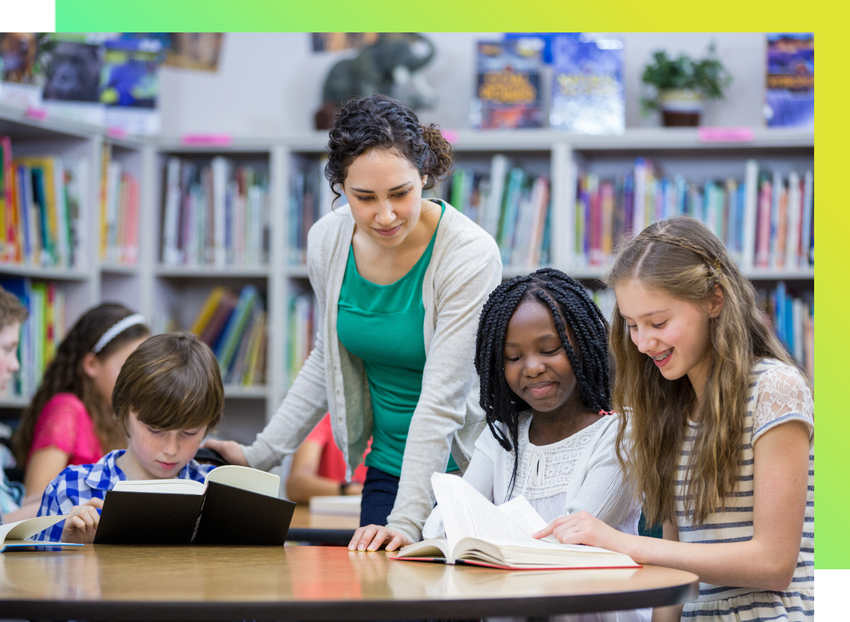 A teacher or educator with dark curly hair, wearing a green shirt and white cardigan, standing beside a table in a school library. 
