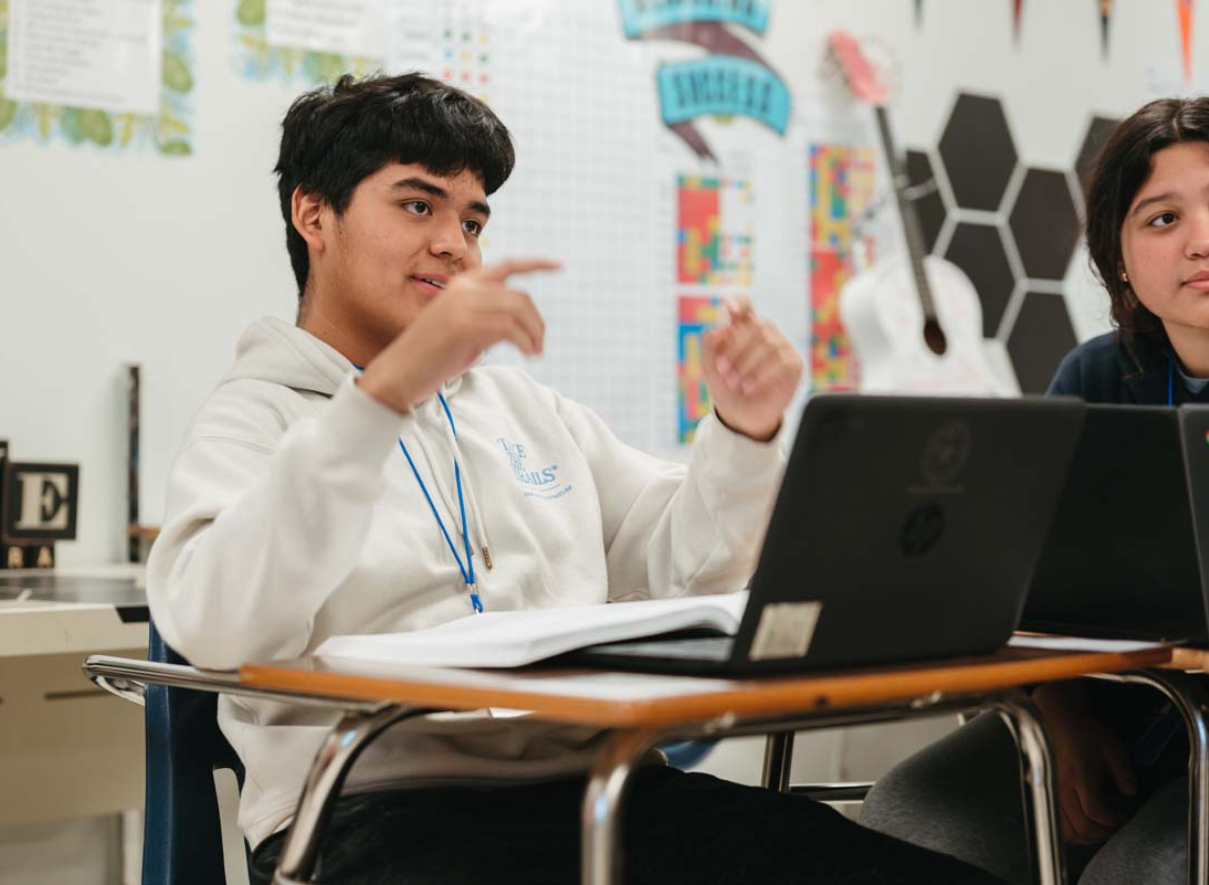Young person with short dark hair wearing a white shirt, sitting at a desk with a laptop. They appear to be in a classroom or study environment with blurred background elements
