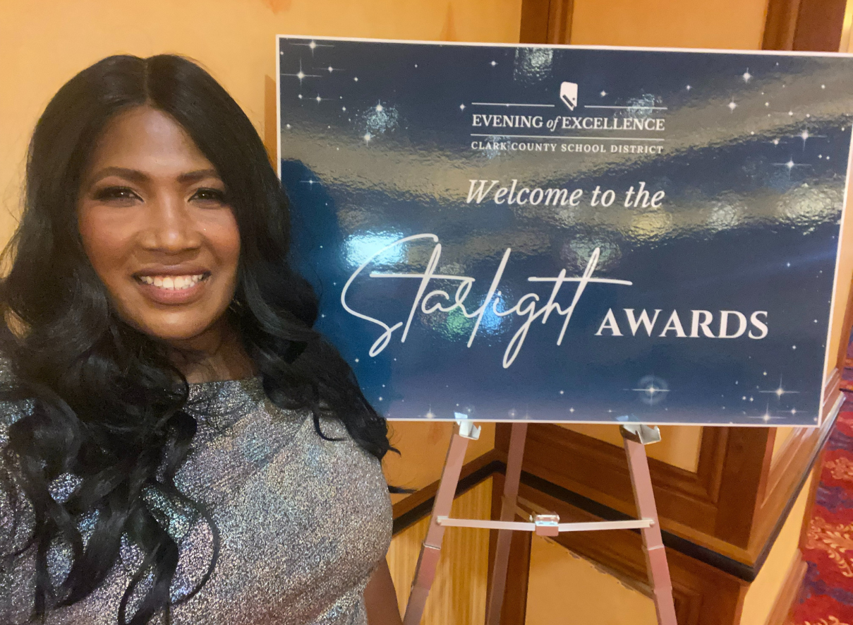 A woman with long dark hair is smiling at the camera. She is standing next to an event sign on an easel that reads 'Welcome to the Spotlight Awards' with 'Evening of Excellence' visible at the top of the sign. 