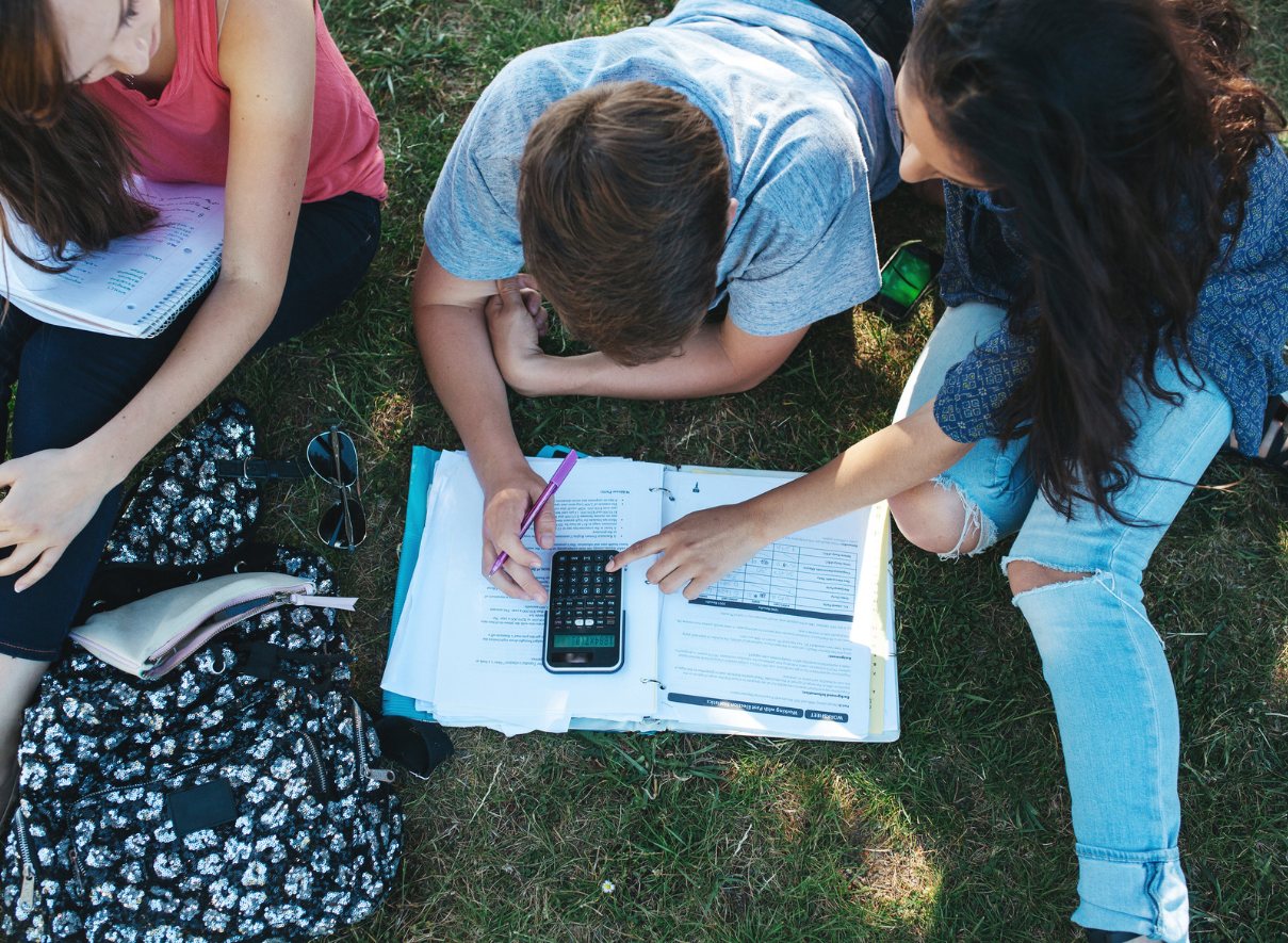Three students studying together outdoors on grass. One person in a light blue t-shirt is leaning over an open notebook while using a calculator. Two others - one wearing pink and another in casual clothing - are sitting nearby with their own materials spread out on the ground.
