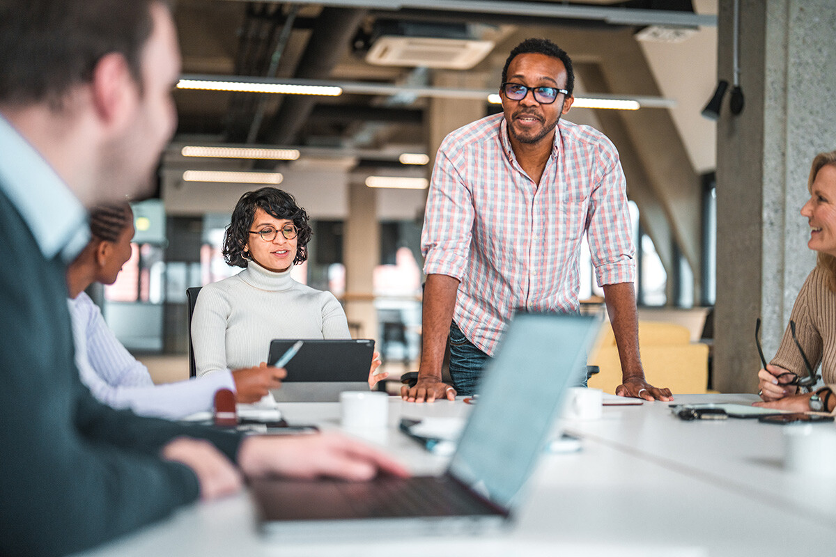 A diverse group of professionals in a modern office meeting. A person wearing glasses and a checkered shirt is standing at the head of a conference table, appearing to lead the discussion.