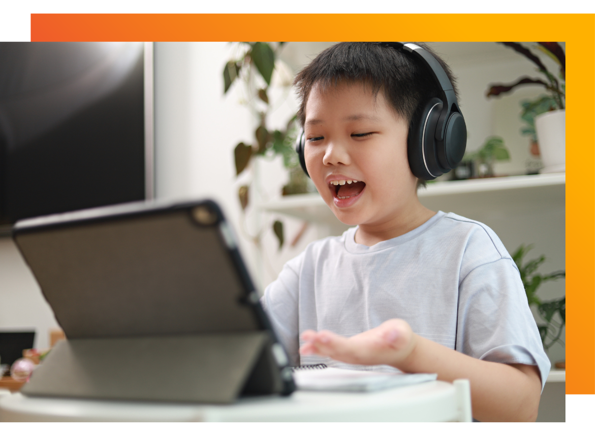 A young Asian boy wearing headphones and a light gray t-shirt, sitting at a table and gesturing with his hands while looking at a tablet device. He appears to be engaged in a video call, online learning session, or interactive digital activity. 