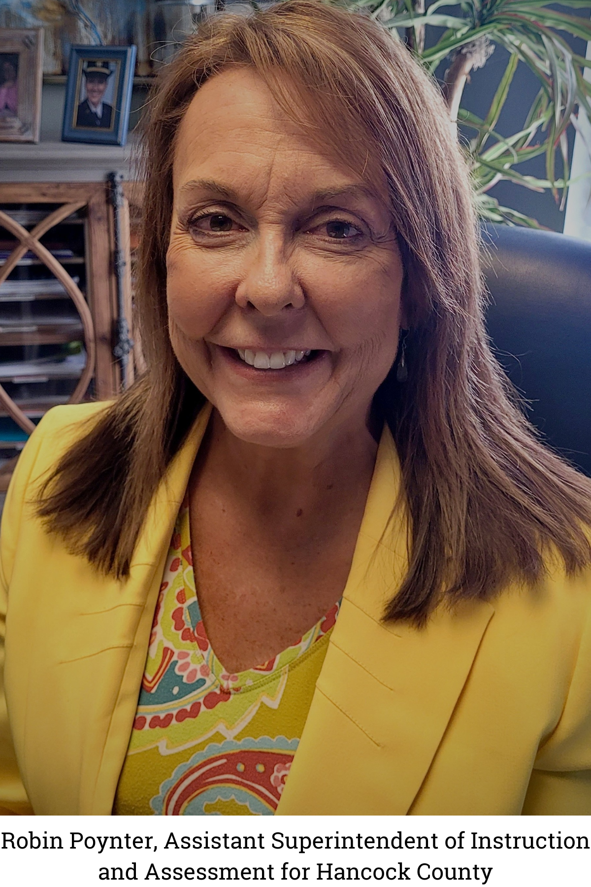 Robin Poynter smiles into the camera wearing a yellow jacket. She is sitting in a black chair and there is a plant behind her.
