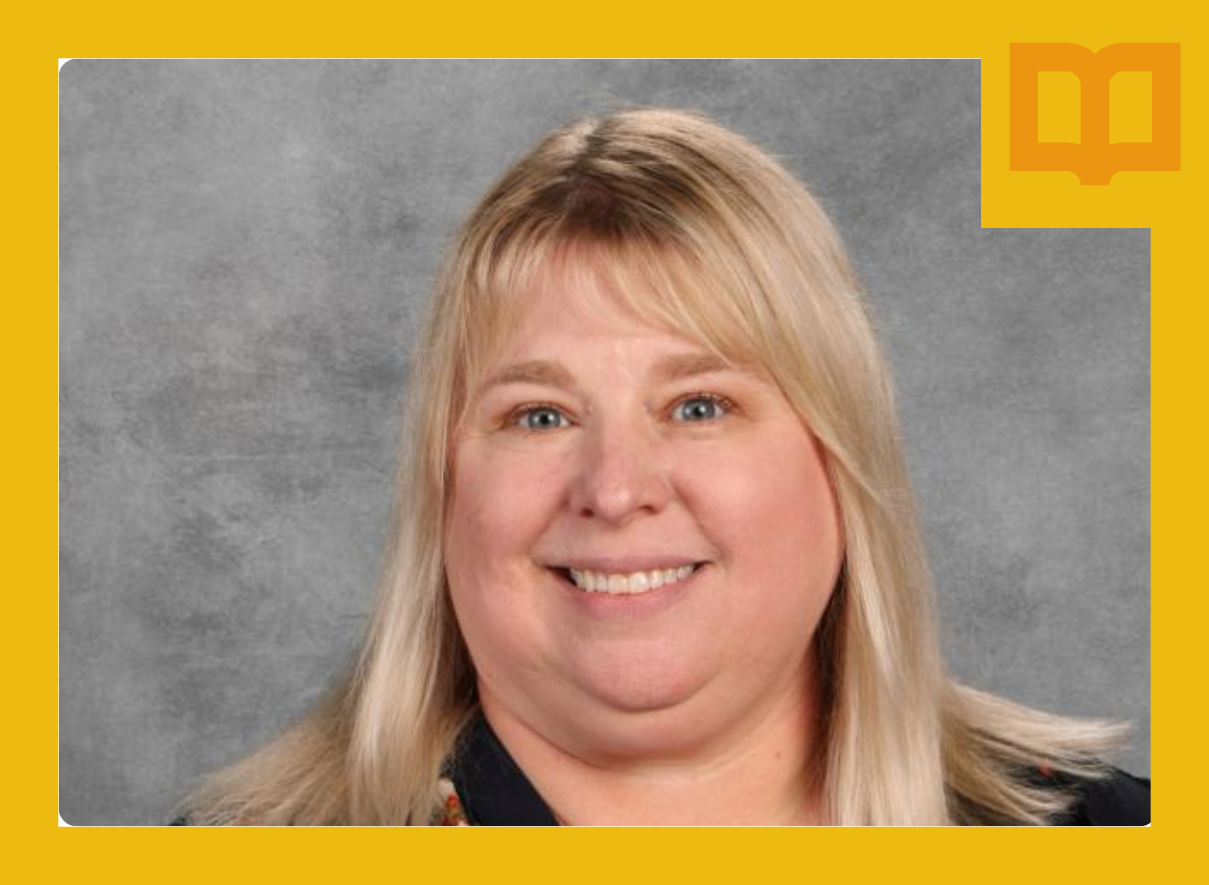 A portrait photograph of a person with blonde hair and bangs, smiling at the camera. They are wearing a dark floral-patterned top against a neutral gray background.