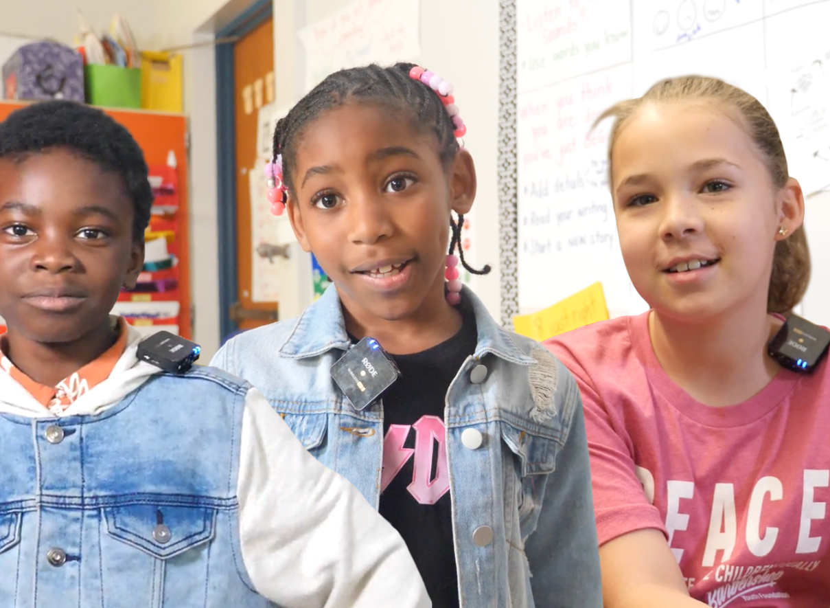 Three young students standing side by side in a classroom, each wearing small microphones or recording devices clipped to their clothing. The classroom has educational materials visible on walls in the background. 