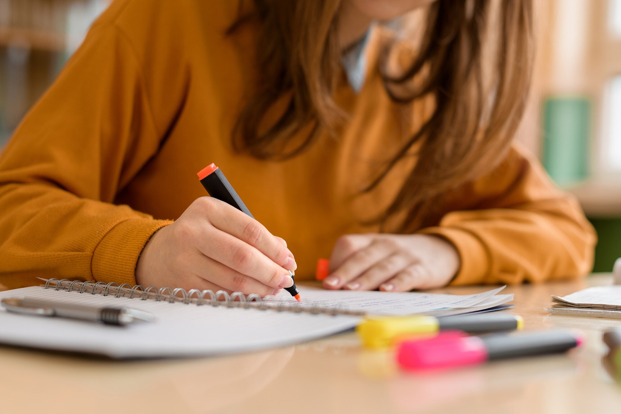 A person with long hair wearing an orange/rust-colored sweater is writing in a notebook at a desk. Their hands are visible as they hold a pen, and there appear to be office supplies including highlighters visible on the work surface.