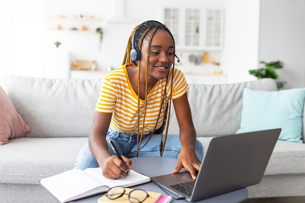 A young Black woman with braided hair working on a laptop while sitting on a couch. She's wearing a yellow and white striped shirt.