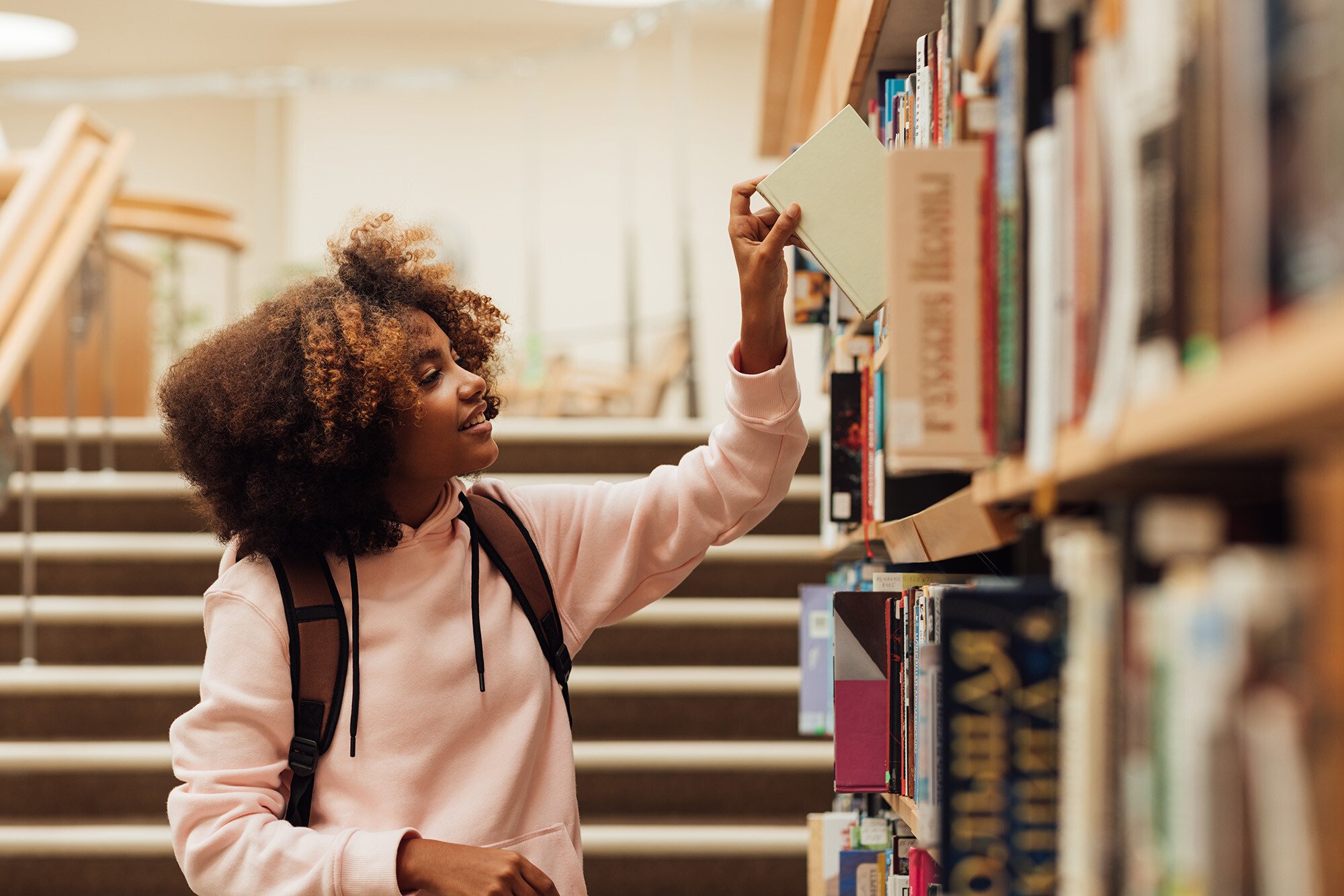 A person with curly hair in a light pink sweater reaches for a book on a library shelf. They appear to be in a library with stairs visible in the background and multiple bookshelves in the foreground. The individual is standing sideways to the camera while selecting a book.