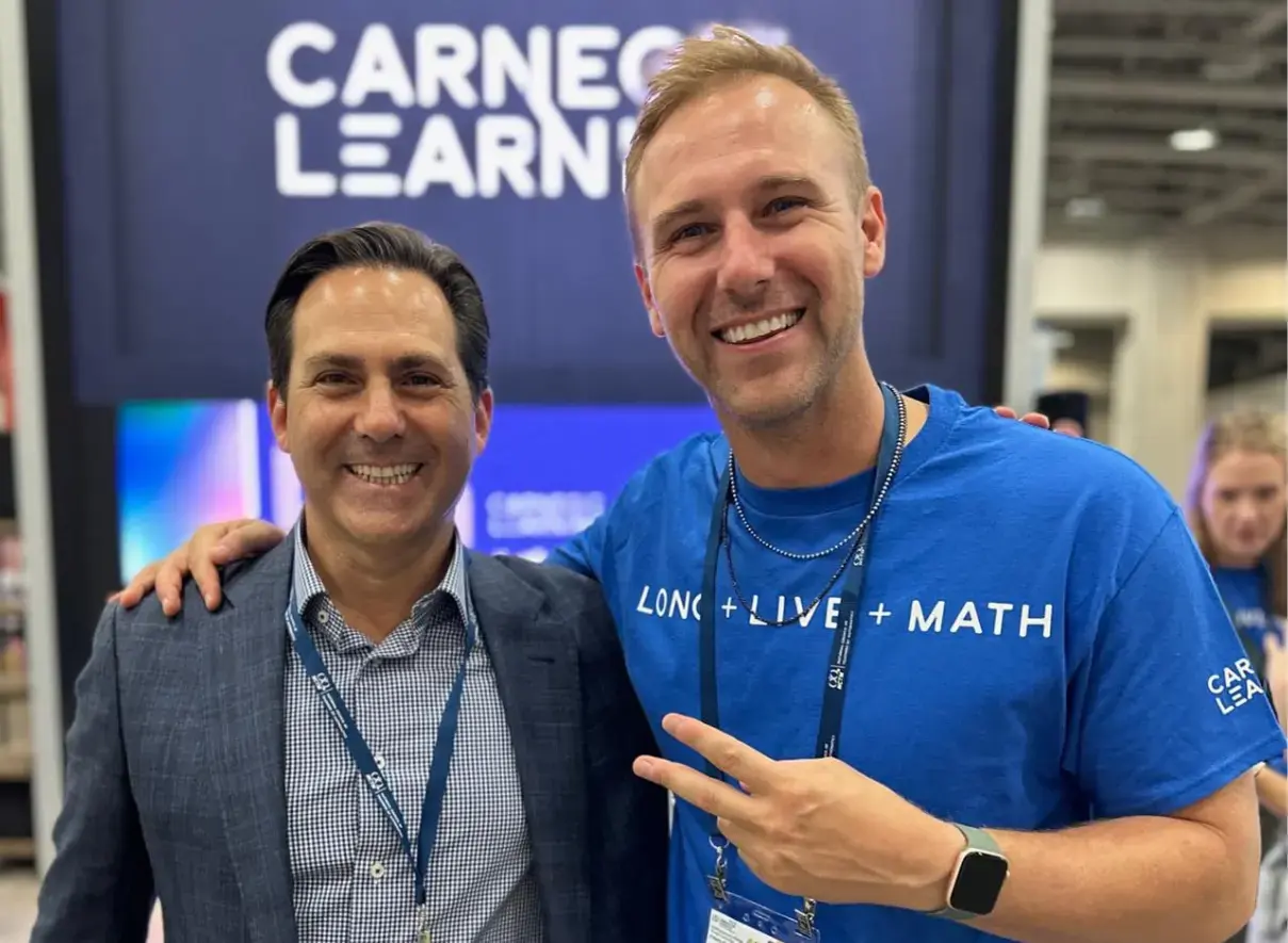 Two men standing together smiling at the camera in what appears to be an event or conference space. One man is wearing a t-shirt with 'MATH' visible on it, and there's a partial 'CARNEGIE LEARN' sign visible in the upper left.