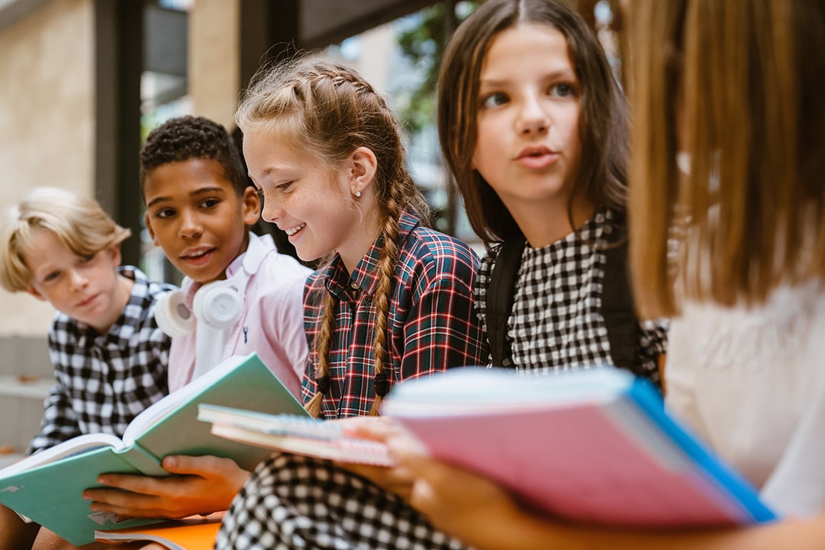A group of four children sit together outside, smiling and talking while holding open books. One child has headphones around his neck. They appear to be enjoying a discussion or study session.