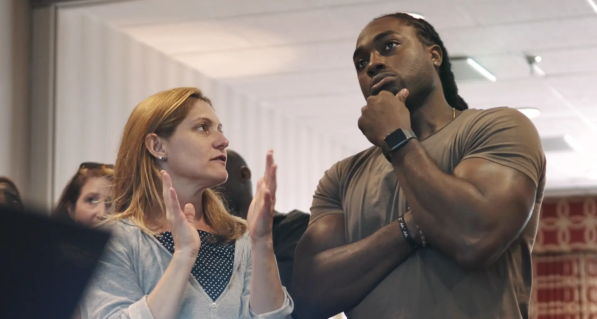 A woman with long brown hair gestures animatedly as she discusses Carnegie Learning with a man sporting braided hair and a smartwatch. He listens intently, hand on chin, in a brightly lit room bustling with several other people.