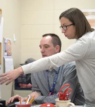 A woman with glasses and a light-colored cardigan leaning over to assist a male colleague who is seated at a desk. She is pointing at something while he appears to be working with office supplies including red scissors visible on the desk.
