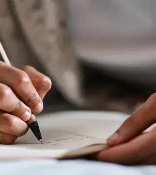 Close-up shot of hands writing on a piece of paper. One hand is holding a pen or pencil while the other hand is steadying the paper, showing the intimate details of writing or note-taking.