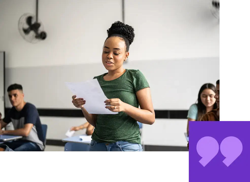 A young woman with her hair in a bun, wearing a green t-shirt and jeans, standing in a classroom while reading from a paper in her hands. Other students can be seen seated in the background. The image has a purple quotation mark graphic in the bottom right corner.